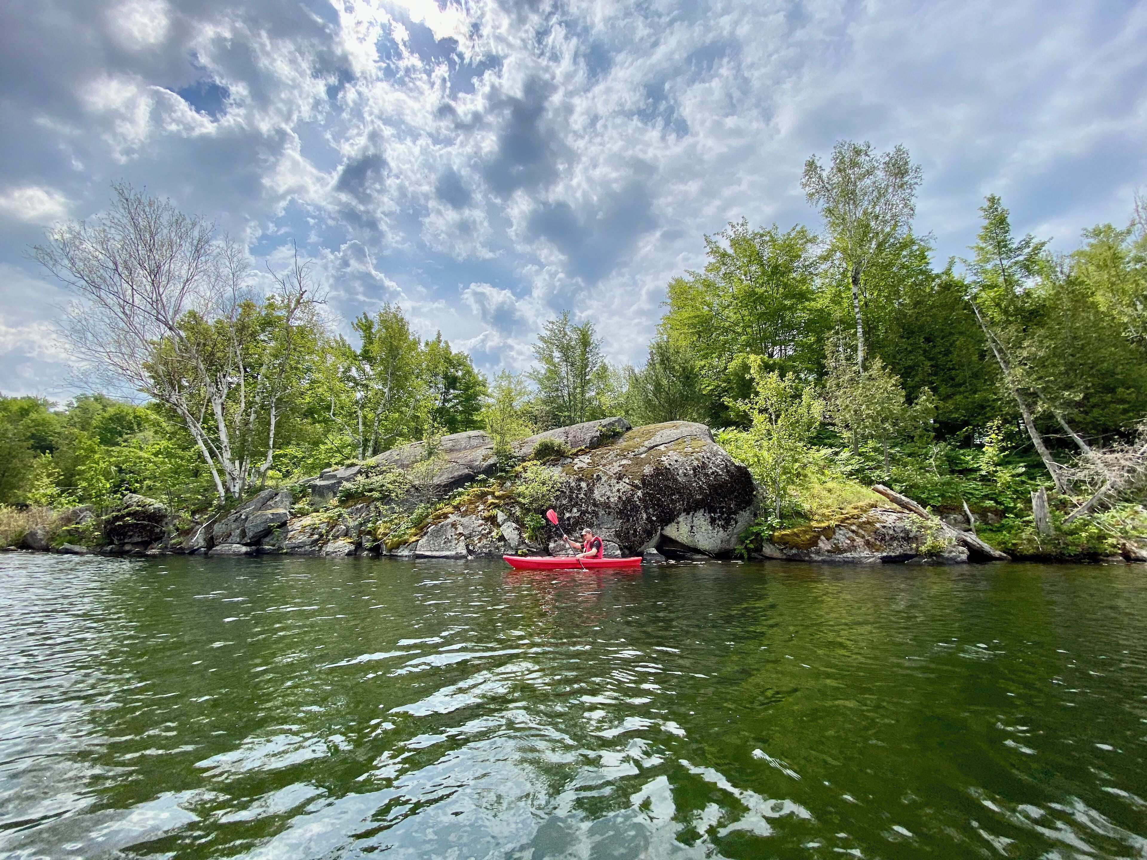 Un kayakiste dans un kayak rouge, sur une rivière paisible, entouré de rochers et d’arbres.