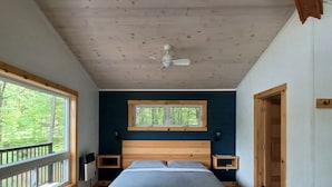 Une chambre avec un lit en bois, une fenêtre donnant sur des arbres, un ventilateur de plafond et un banc en bois.