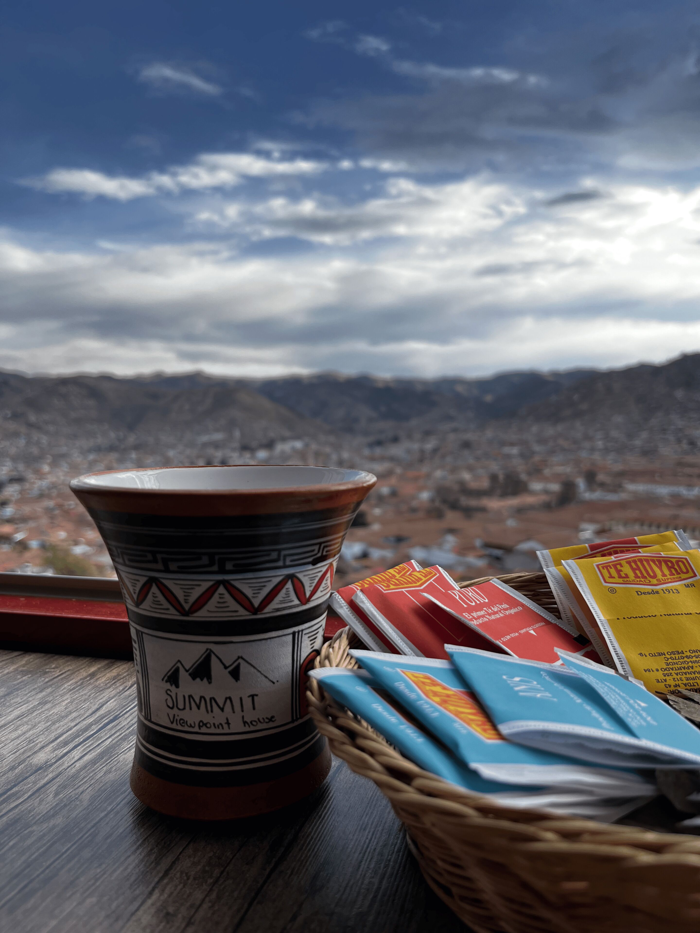 A terracotta mug with a mountain design, a basket of books, and a view of a cityscape and mountains in the background.