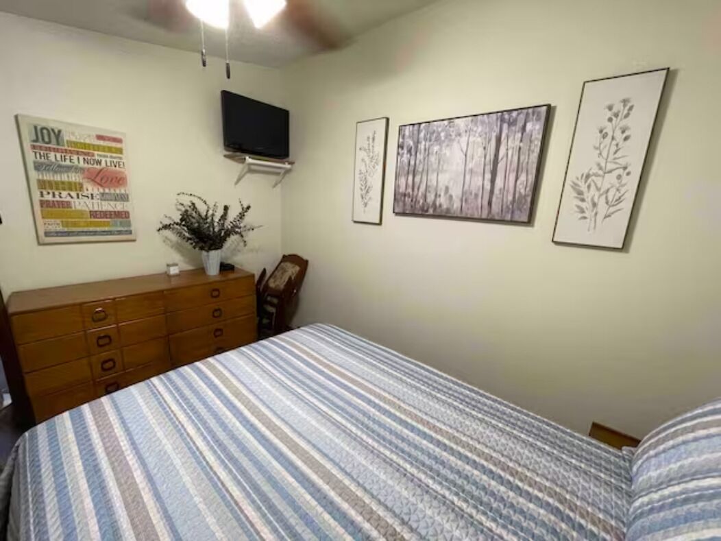 Un dormitorio con cama, cómoda, televisor de pantalla plana en la pared y cuadros enmarcados.