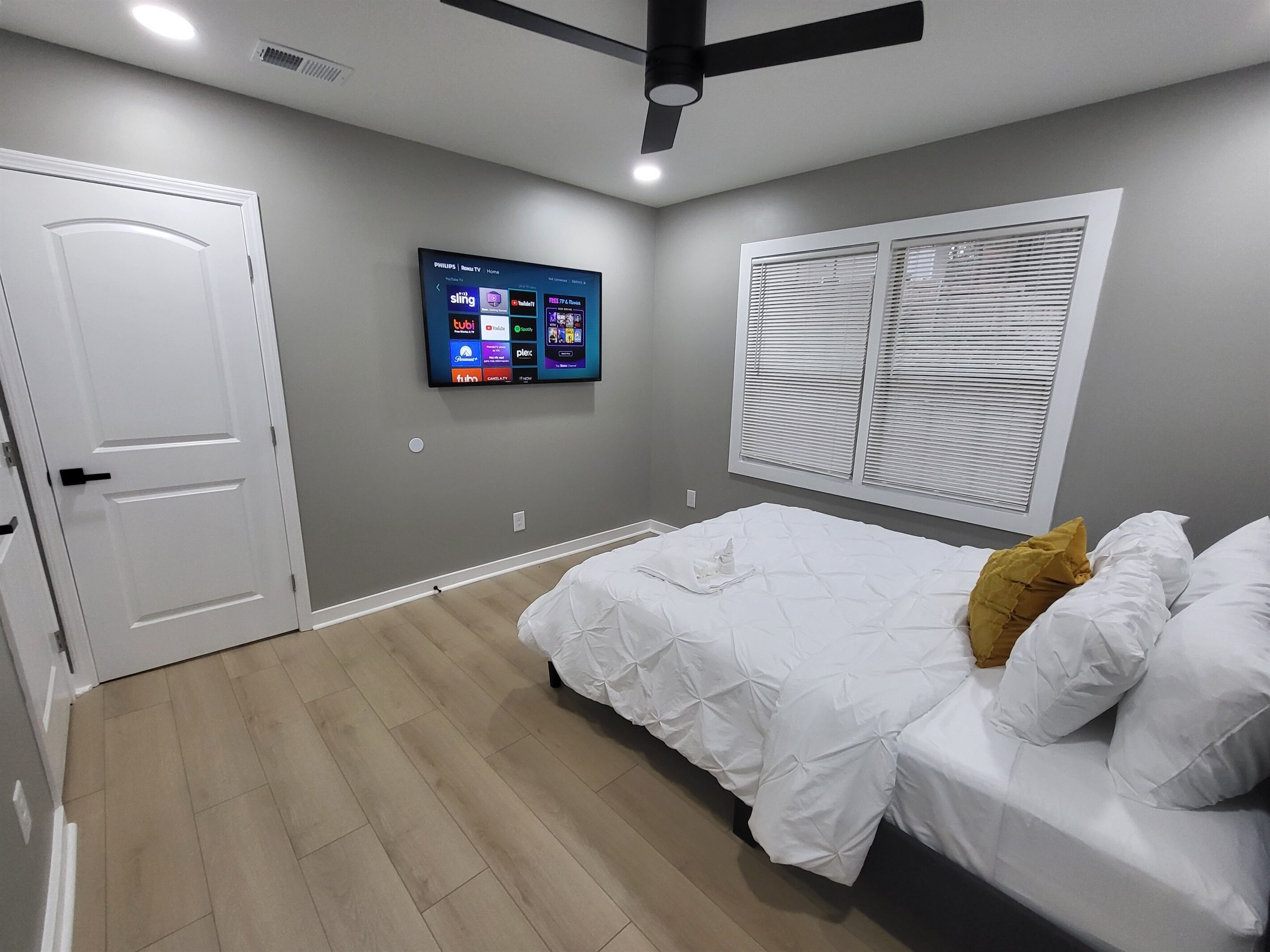 A bedroom with a bed, a TV mounted on the wall, a ceiling fan, and a window with blinds.