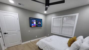 A bedroom with a bed, a TV mounted on the wall, a ceiling fan, and a window with blinds.