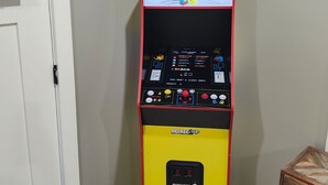 En Pac-Man-spelautomat i ett rum med en vit dörr, ett träbord och en vägg dekorerad med Pac-Man- och spökfigurer.