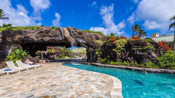 Uma área de piscina com um deck de pedra, uma cascata e vegetação tropical exuberante.