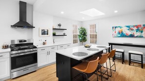 A modern kitchen with white cabinetry, a stainless steel stove, and a black range hood. There is a large island with stools and a colorful abstract painting on the wall. - Clean, Classy, Convenient. Montclair's Best Stay! Walk to town and train (Montclair)