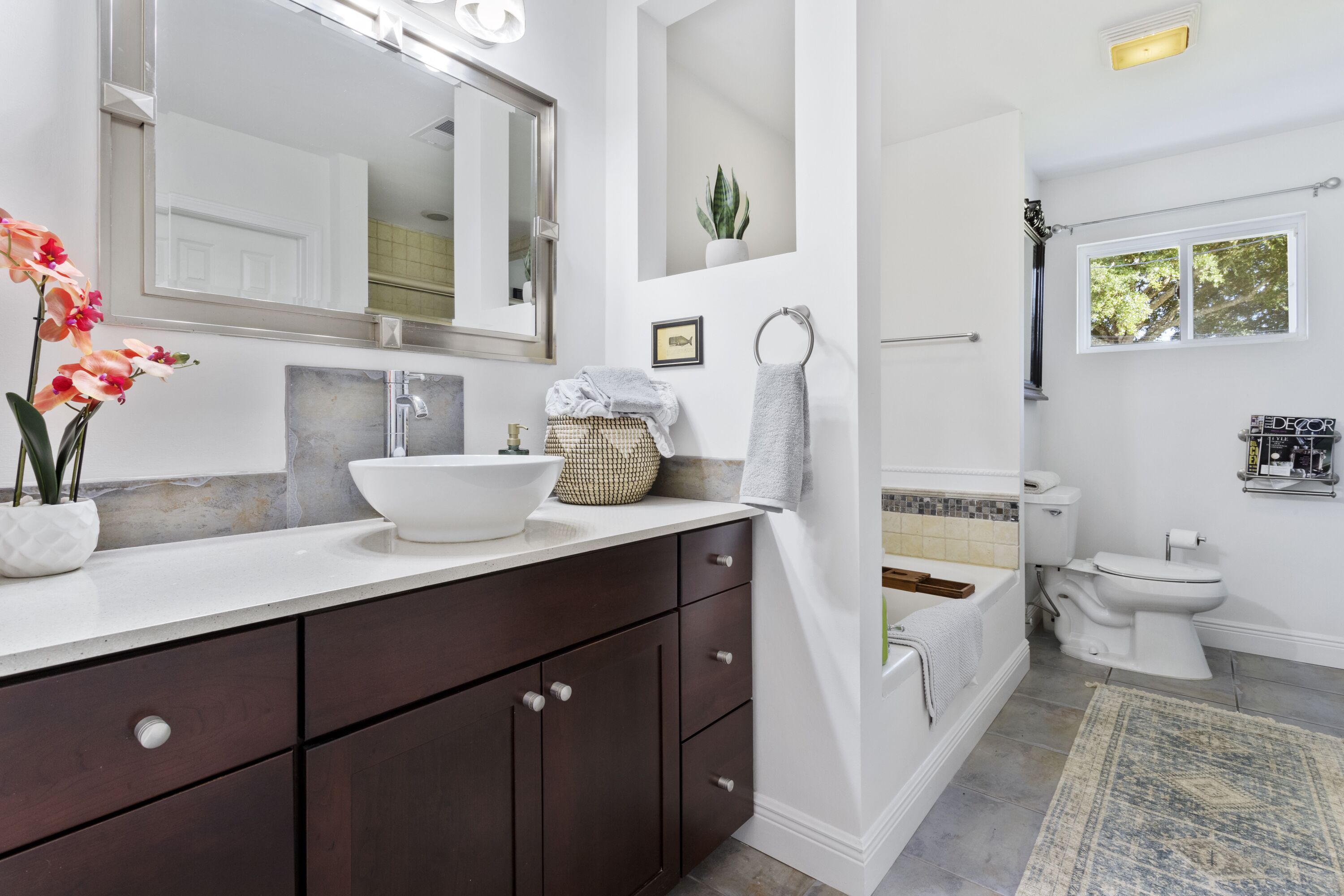 A modern bathroom with a large mirror, white sink, dark wooden cabinets, a bathtub, and a toilet.