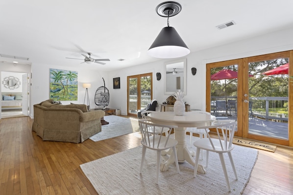 Ein geräumiges Wohnzimmer mit Holzboden, einem beigefarbenen Sofa, einem Esstisch mit weißen Stühlen, einem Deckenventilator und einem großen Fenster mit einem roten Regenschirm.
