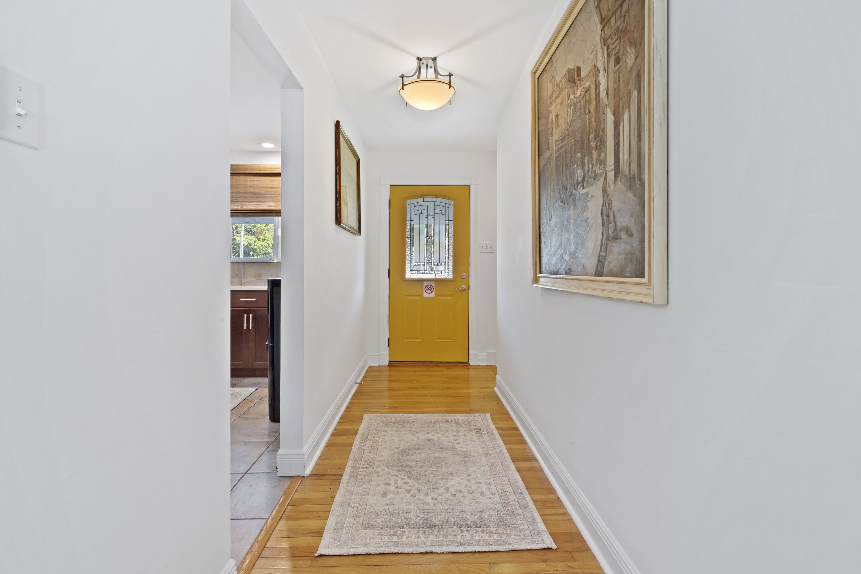 A hallway with a wooden floor, a rug, a yellow door, and a painting on the wall.