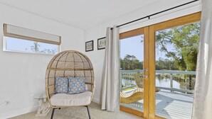 A room with a wicker chair, a glass table, a view of a lake, and a wooden deck.