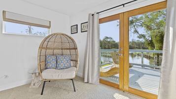 A room with a wicker chair, a glass table, a view of a lake, and a wooden deck.