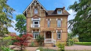 Uma casa grande de dois andares com um telhado em galbe e várias janelas, e um pequeno alpendre com guarda-corpo.