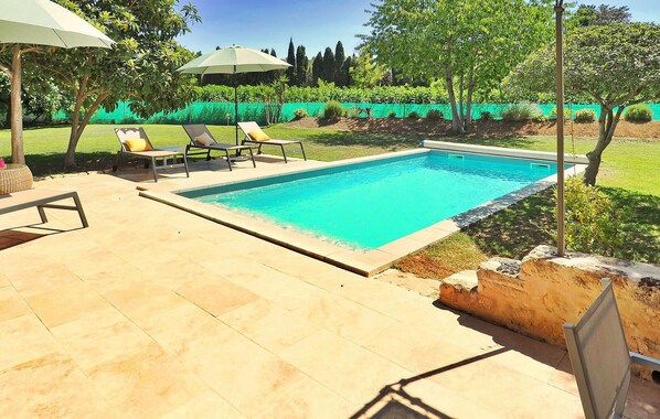 Piscina retangular com deck ladrilhado, rodeada por um hedge verde e árvores. Há espreguiçadeiras e um guarda-sol.