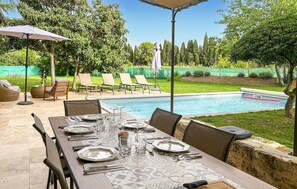 Uma área de refeições ao ar livre com piscina, espreguiçadeiras e uma mesa coberta.