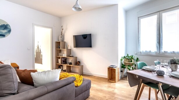 A modern living room with a grey sofa, wooden floor, a TV mounted on the wall, and a dining area with a table and chairs.