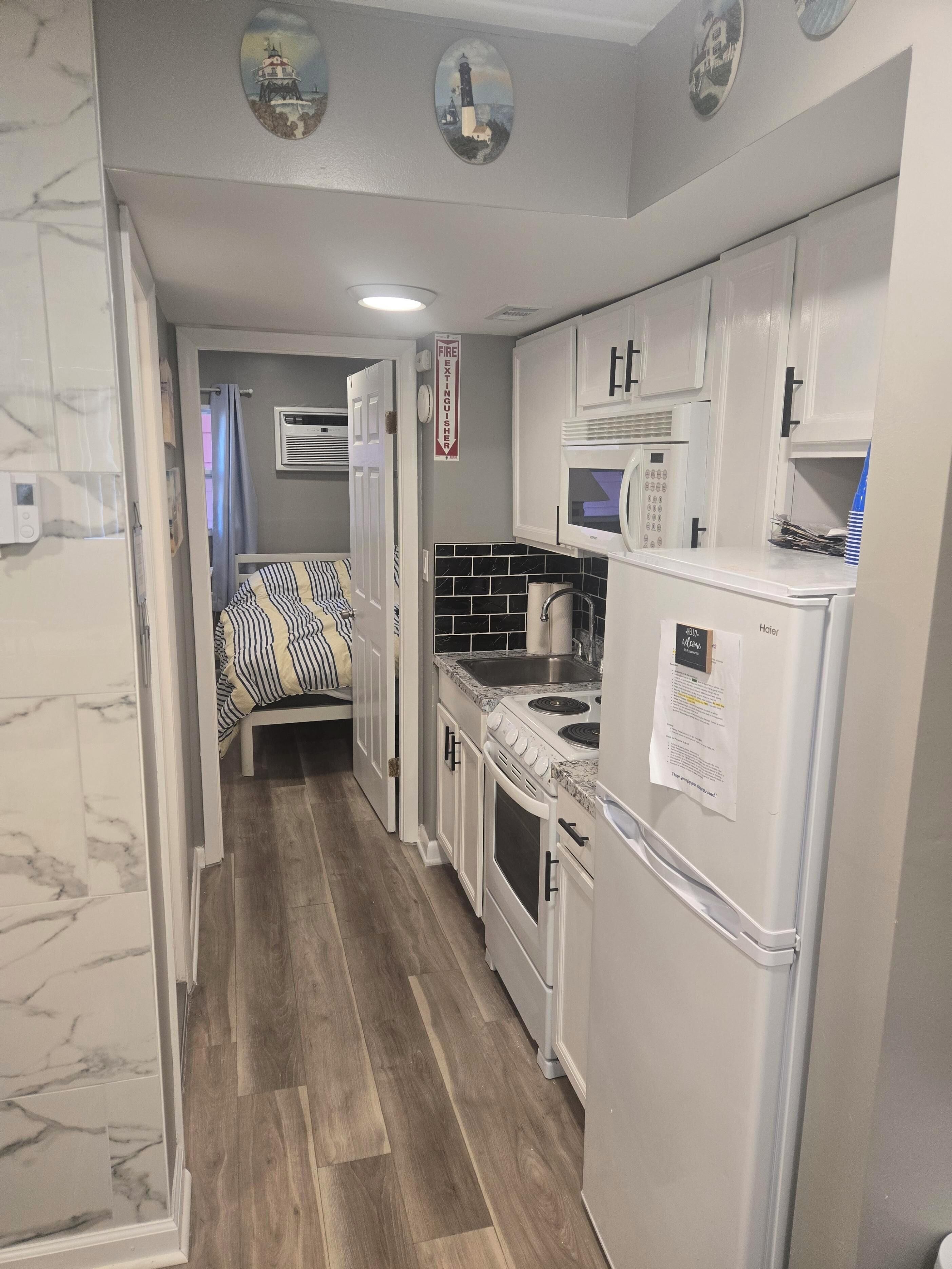 A compact kitchen with white cabinets, a microwave, a stove, and a sink. There is a bed with striped bedding in the adjacent room.