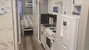 Une cuisine compacte avec des meubles blancs, un four à micro-ondes, une cuisinière et un évier. La pièce adjacente contient un lit avec une literie à rayures.