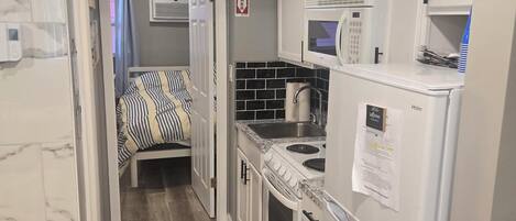A compact kitchen with white cabinets, a microwave, a stove, and a sink. There is a bed with striped bedding in the adjacent room.