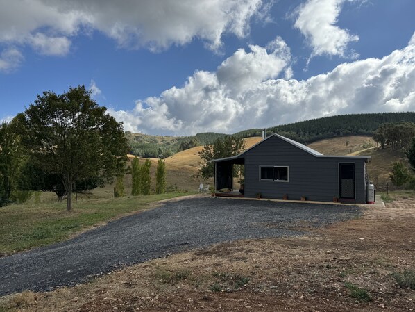 A small, dark-colored cabin with a gravel driveway and a large tree to the left. - Native Valley Cottage is a peaceful, tranquil hideaway in the Tumbarumba area. (Carabost)