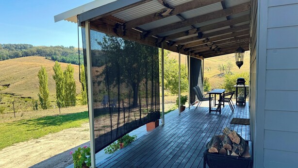 Une terrasse extérieure couverte, dotée d’une table et de chaises, d’un foyer et offrant une vue sur un paysage vallonné.