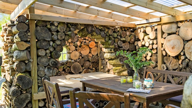 Un comedor al aire libre de madera con una mesa y sillas, rodeado por un muro de troncos apilados.