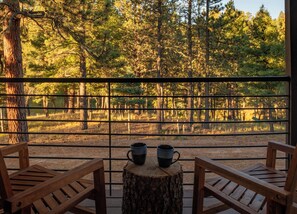 A wooden deck with two chairs and a table, overlooking a forested area through a metal railing.
