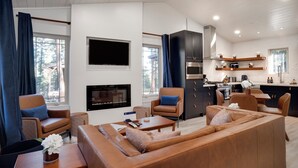 A modern living room with a fireplace, leather sofas, and a kitchen area with dark cabinets and a stove. - Incredible Cabin, Minutes to the Resort, King Bed, A/C, Fire pit (Angel Fire)