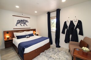 A modern bedroom with a large bed, a wooden headboard, a nightstand with a lamp, a fluffy rug, a window with curtains, and a seating area with a chair and a table. - Incredible Cabin, Minutes to the Resort, King Bed, A/C, Fire pit (Angel Fire)