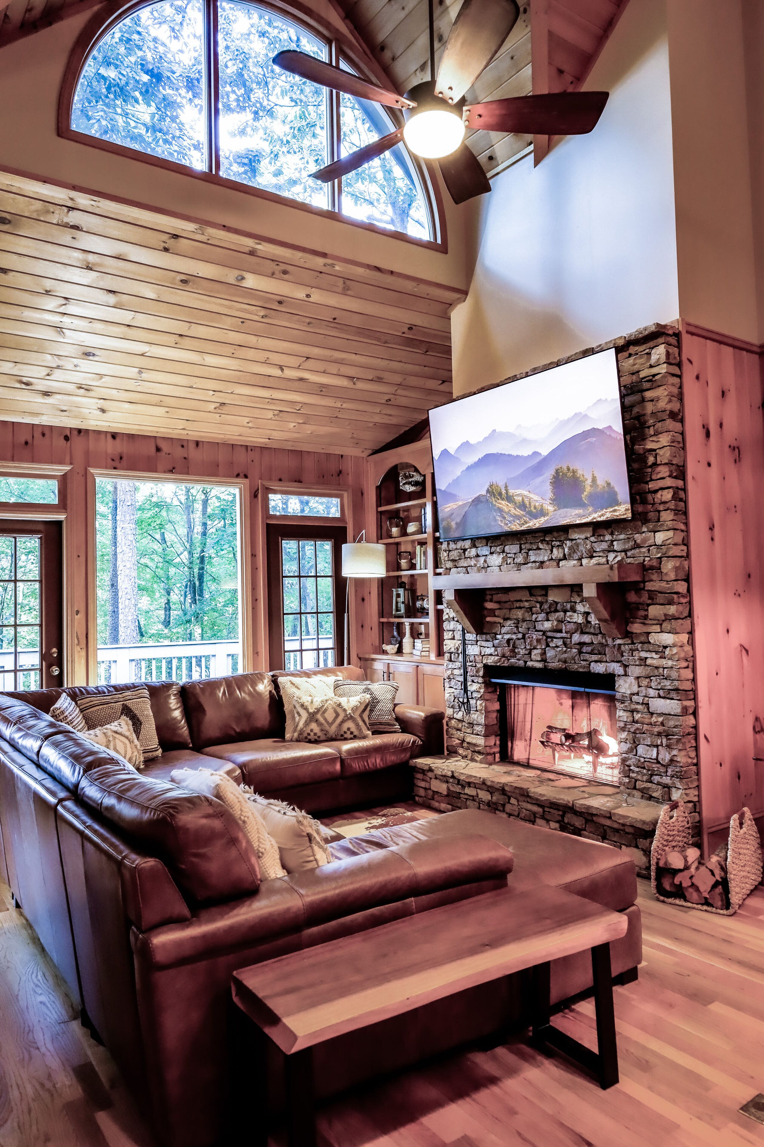A cozy cabin interior with a stone fireplace, leather sofa, wooden ceiling, and large windows.