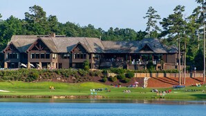 A large, multi-story lodge with a prominent porch and multiple windows, situated near a lake with a well-maintained lawn and a golf course in the background. - Discover family fun and serenity at Big Canoe resort! (Jasper, GA)
