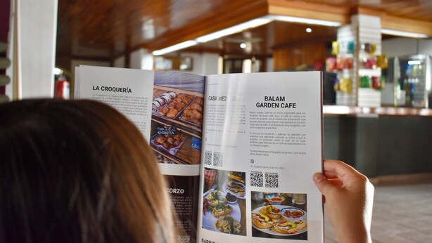 A person reading a brochure with sections titled "LA CROQUERÍA" and "BALAM GARDEN CAFE". - Hotel Garden Inn  (Aguascalientes)