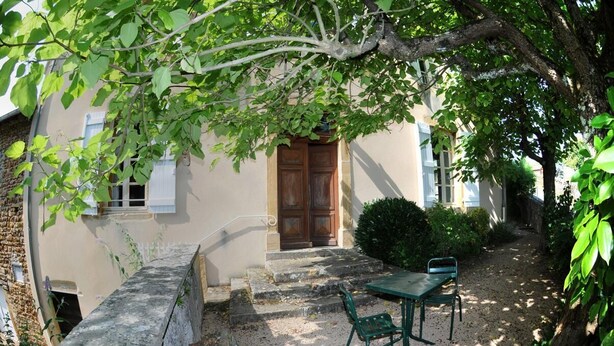 A courtyard with a stone bench, a table, and chairs, surrounded by greenery and a building with a wooden door.