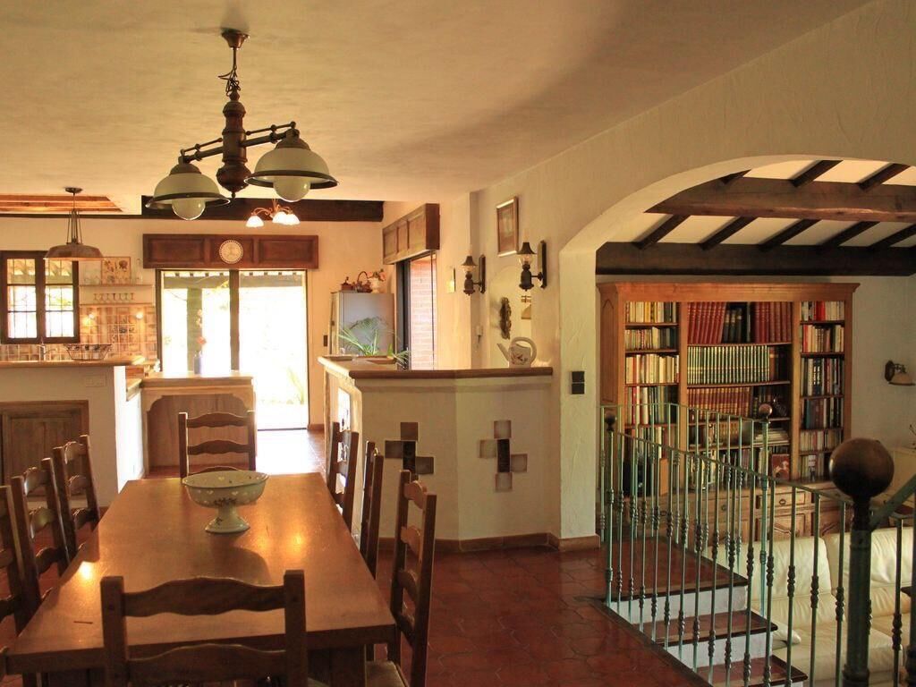 A dining area with a wooden table and chairs, a chandelier, and a staircase leading to a library with bookshelves.