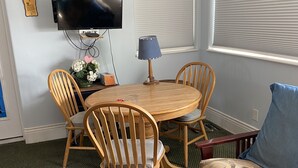 Ein Zimmer mit einem runden Holztisch, vier Holzstühlen, einem an der Wand befestigten Fernseher, einer Lampe auf dem Tisch und einem Sofa mit einem gemusterten Kissen.