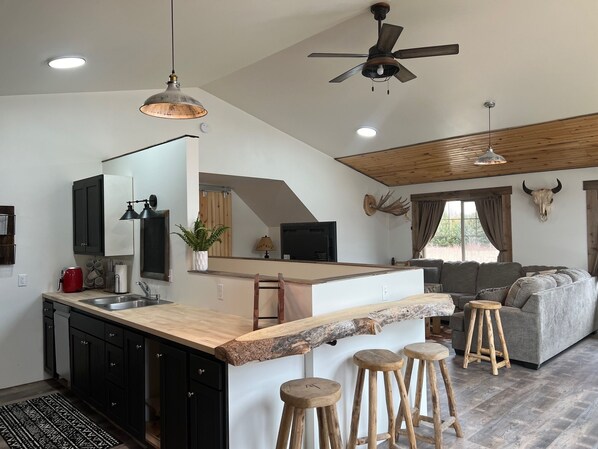 Una cocina moderna con una isla central, taburetes de bar, un ventilador de techo y una zona de estar con sofá y una calavera de animal montada.