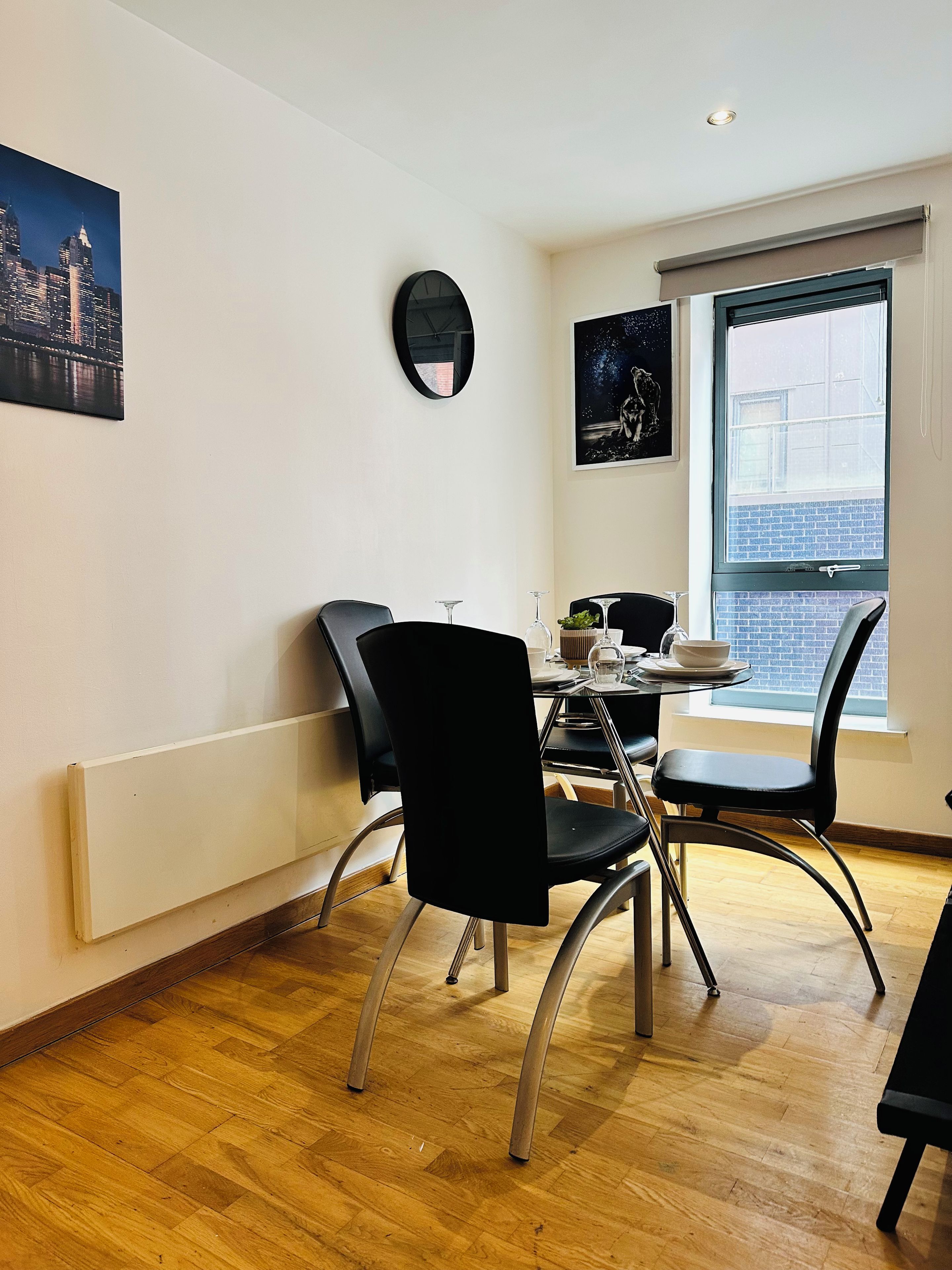 A dining area with a table set for two, two black chairs, a round mirror on the wall, and a window with a view of buildings.