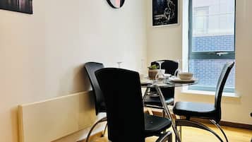A dining area with a table set for two, two black chairs, a round mirror on the wall, and a window with a view of buildings.