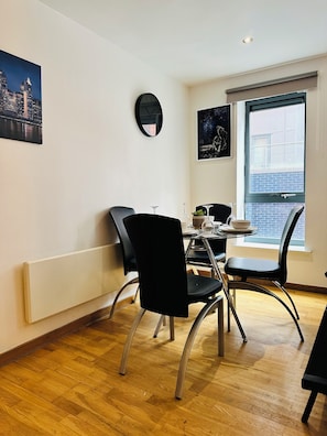 A dining area with a table set for two, two black chairs, a round mirror on the wall, and a window with a view of buildings. - Luxury 2 Bedroom Apartment in City Centre  (Liverpool)