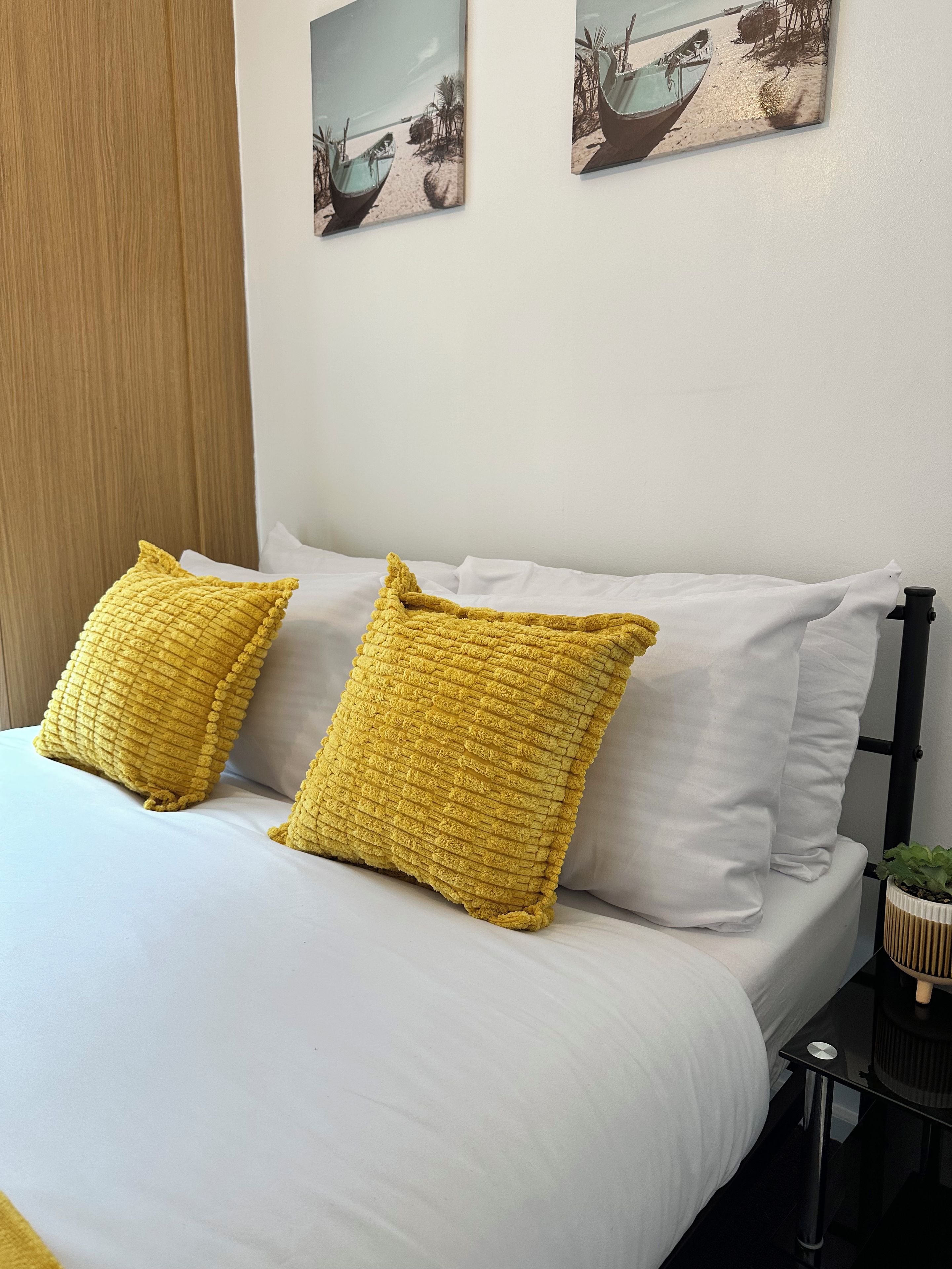A bed with white bedding and two yellow pillows, a wooden door, and two wall-mounted pictures of boats on a beach.