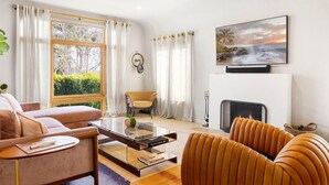 Uma sala de estar moderna com um sofá, poltrona e mesa de centro de vidro. Há uma lareira e uma televisão fixada na parede.