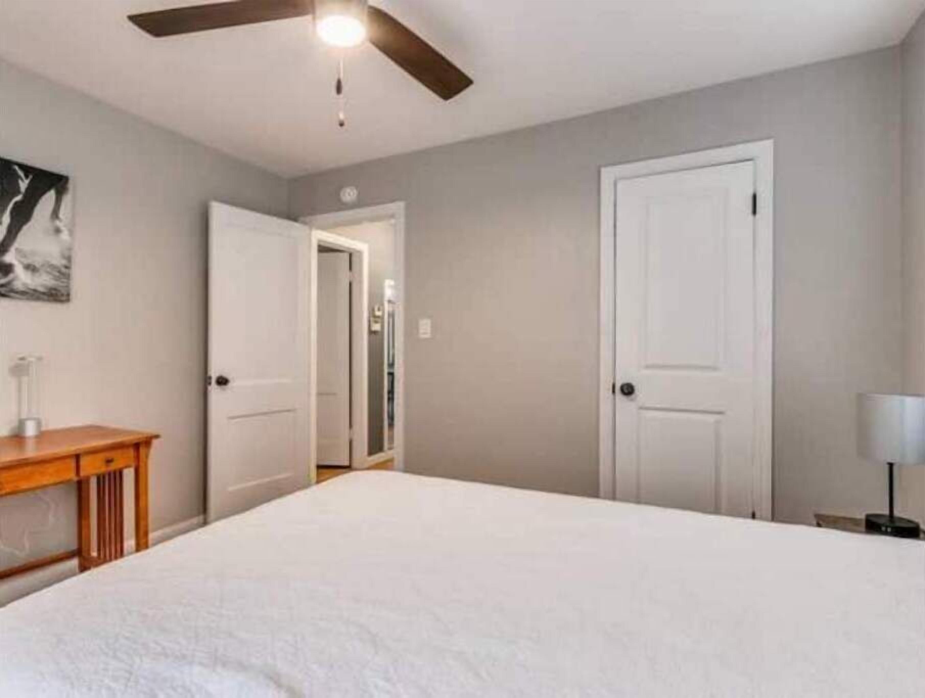 Un dormitorio con una cama, una mesita de noche de madera, un ventilador de techo y una puerta que da a otra habitación.