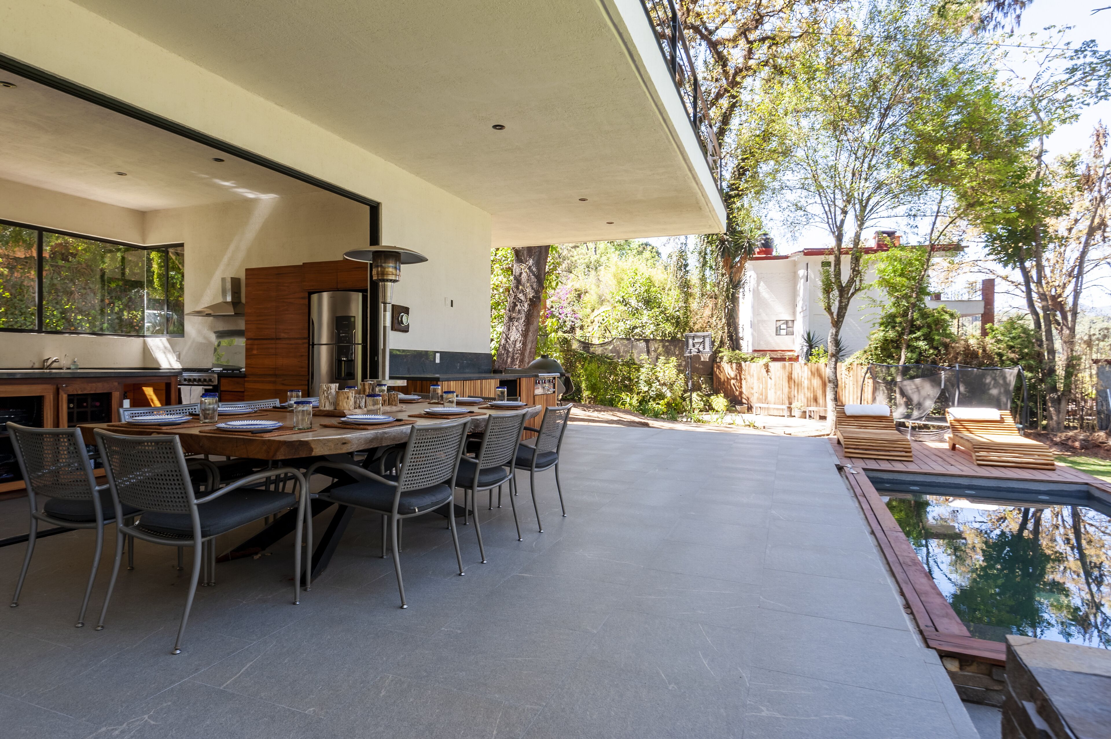 A modern outdoor dining area with a pool, surrounded by trees and a view of a neighboring building.
