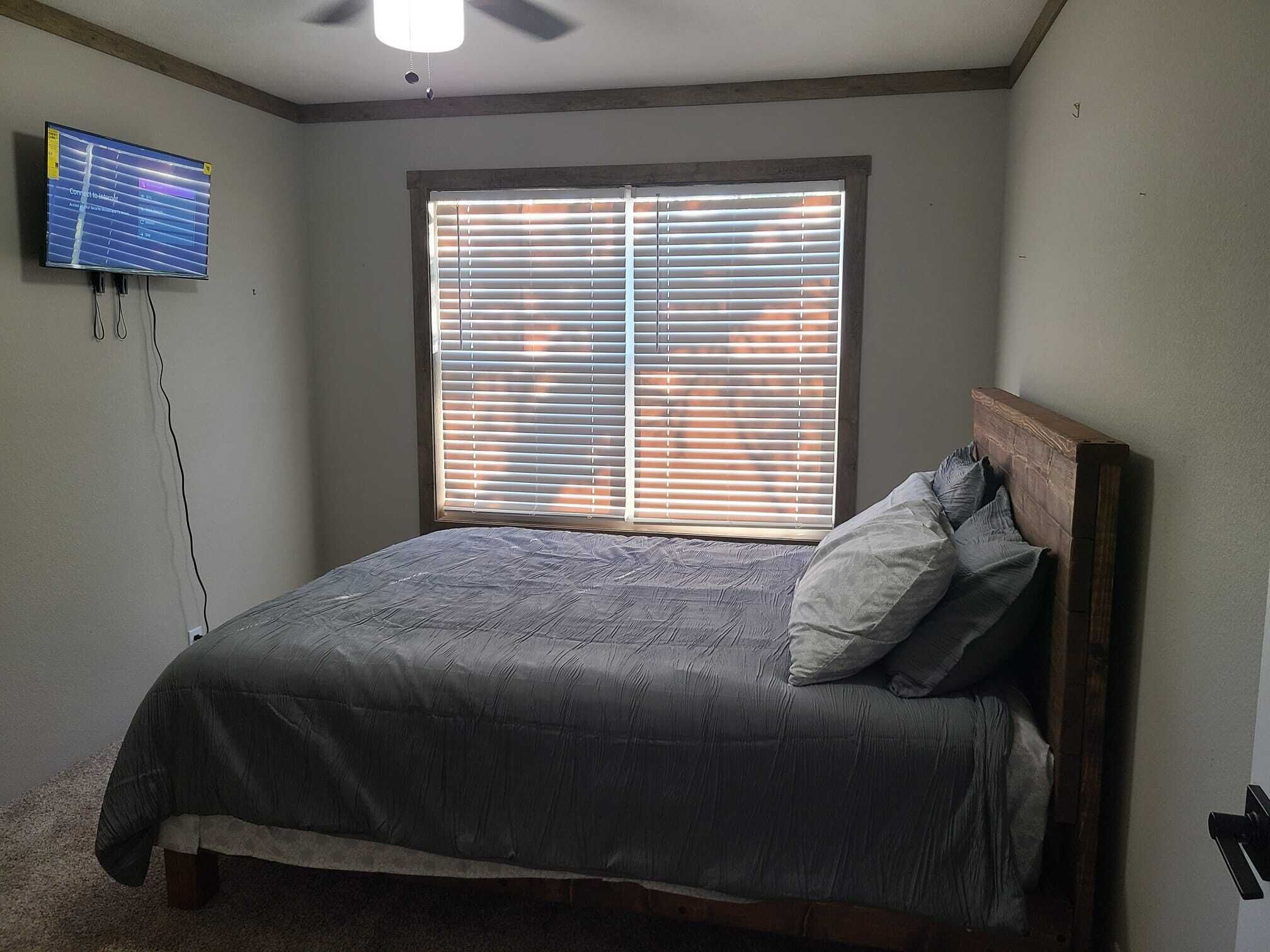 A bedroom with a bed, a window with blinds, a ceiling fan, and a TV mounted on the wall.