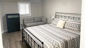 Un dormitorio con una cama, un sofá gris, un archivador negro y una ventana con persianas.