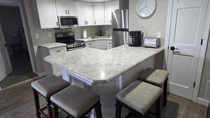 Una cocina con armarios blancos, un refrigerador de acero inoxidable, un microondas, un tostador y un reloj en la pared. Hay cuatro taburetes de bar con asientos acolchados.