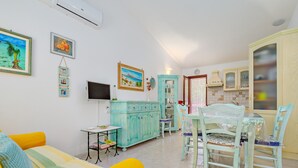 A living room with a sofa, a dining table with chairs, a TV, and a kitchen area with cabinets and a window.