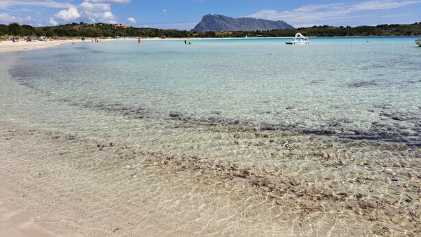 A beach with clear water, a mountain in the background, and a few boats.