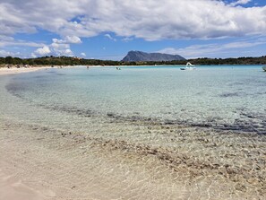 Una playa con agua cristalina, una montaña al fondo y algunos barcos.