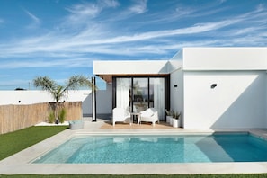 Un espace piscine extérieur moderne, avec un ciel bleu dégagé, un bâtiment blanc et une terrasse en bois.