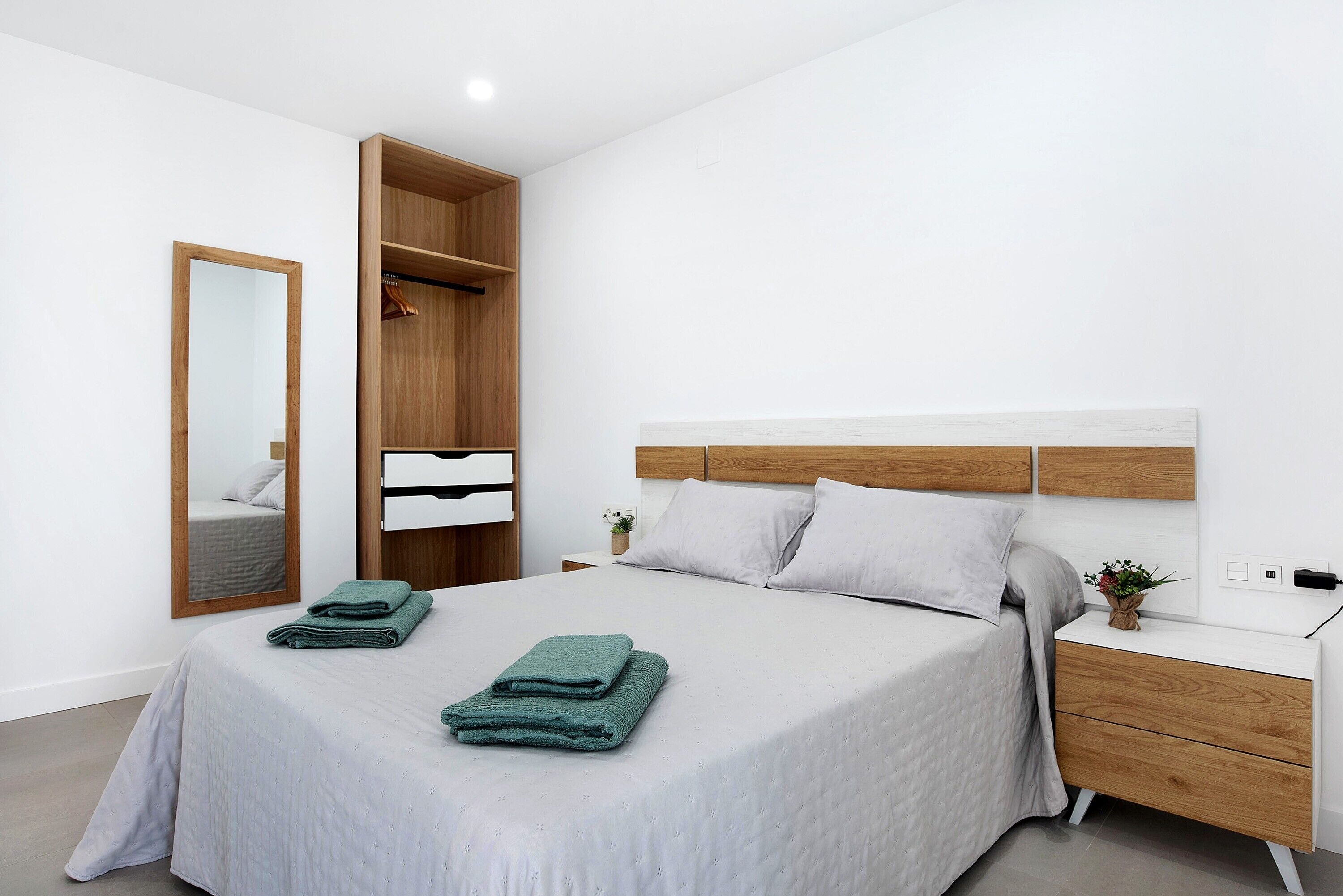 A neatly made bed with a grey bedspread and two folded green towels, a wooden wardrobe with a mirror, and a bedside table with a small plant.