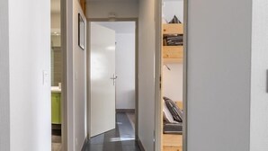Ein schmaler Korridor mit Fliesenboden, einer Holztür am Ende und einem sichtbaren Etagenbett durch eine geöffnete Tür.